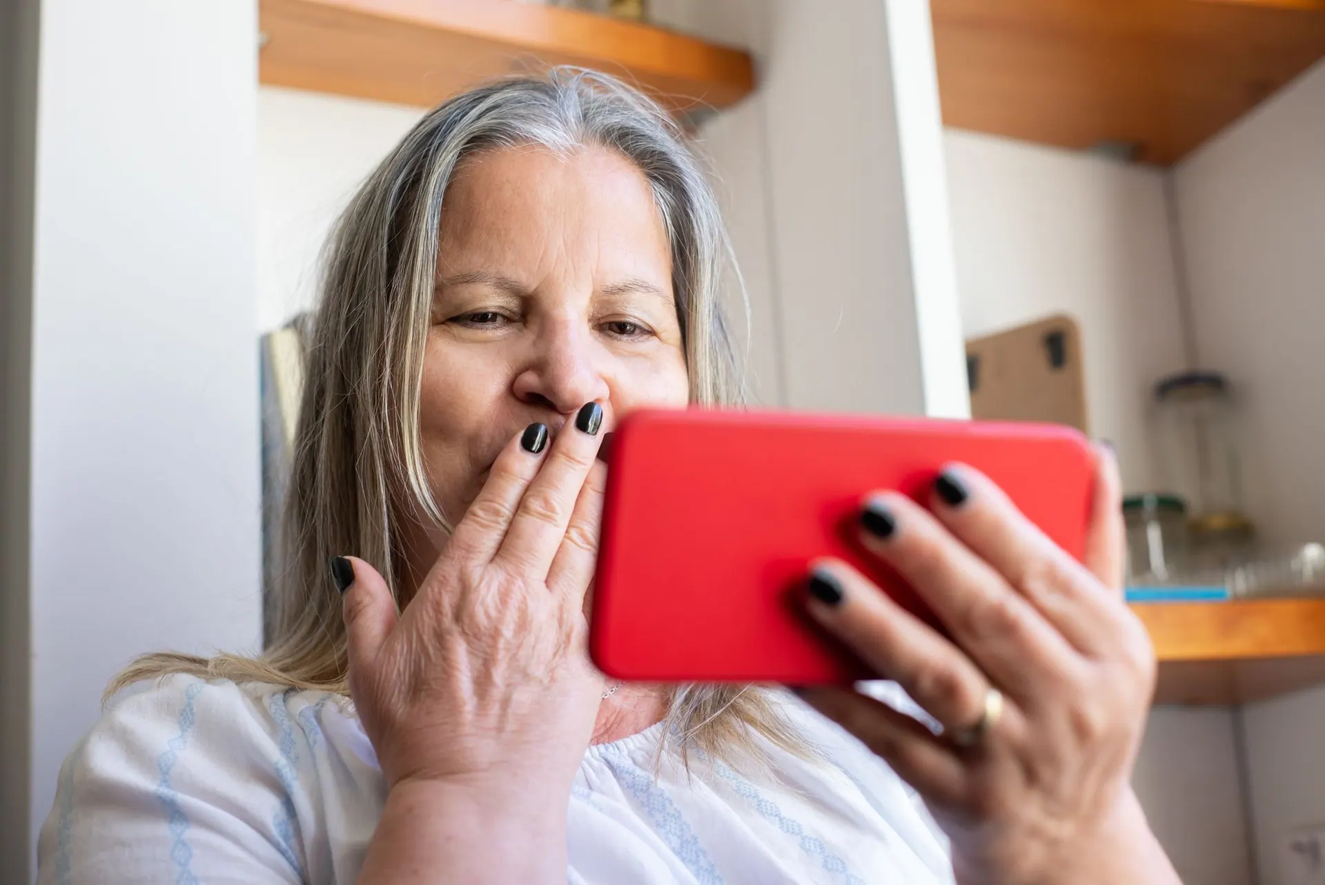 A woman in her sixties covers her mouth in delight as she looks at her phone in the kitchen.